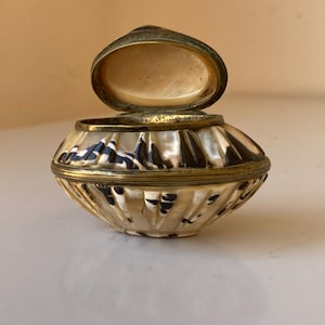 May include: A small, oval-shaped trinket box crafted from iridescent shell with a natural cream and dark brown mottled pattern. The container features a hinged lid with a metallic brass-toned rim and a smooth, light-colored interior lining.