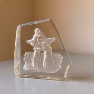 May include: A clear glass paperweight with a white, three-dimensional carving of an inukshuk, a traditional Inuit stone structure.