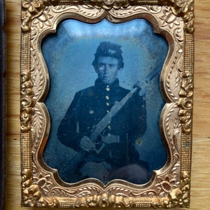 Può includere: Una fotografia in bianco e nero di un uomo in uniforme militare che tiene un fucile. L'uomo indossa un berretto e ha un'espressione seria sul viso. La fotografia è incorniciata in una cornice dorata ornata.