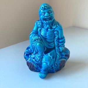 Peut inclure: Une figurine en céramique bleue d'un Bouddha souriant assis sur un rocher. La figurine est détaillée avec des sculptures complexes et une expression sereine.