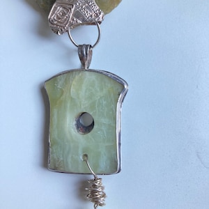 May include: A silver and green jade necklace with a large, rectangular pendant featuring a hole in the center. The pendant is attached to a smaller, round jade bead by a silver wire.