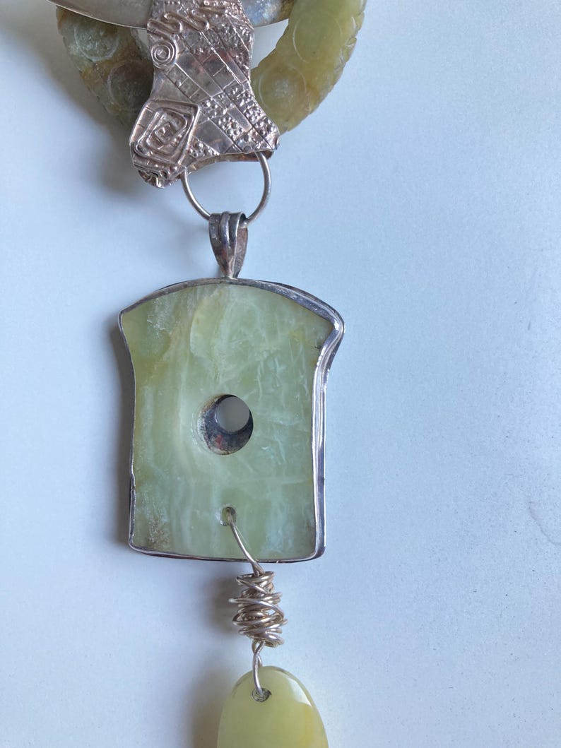 May include: A silver and green jade necklace with a large, rectangular pendant featuring a hole in the center. The pendant is attached to a smaller, round jade bead by a silver wire.