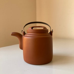 May include: A small, terracotta teapot with a metal handle. The teapot has a rounded body and a spout on the side.