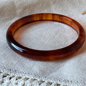 May include: A vintage-style bangle bracelet in shades of amber and brown. The circular bracelet has a smooth, glossy finish and is set against a neutral background. This accessory is a classic piece of jewelry.