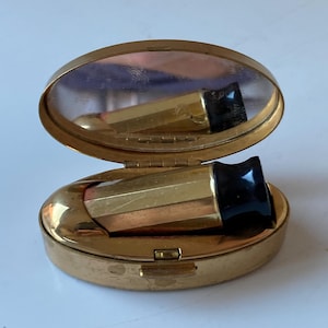 Puede incluir: Un estuche compacto dorado vintage con tapa abisagrada, que revela un pequeño espejo y un lápiz labial. El lápiz labial tiene una funda dorada y una tapa negra. El estuche compacto es de forma ovalada y tiene un cierre de broche.