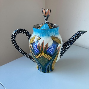 May include: A white ceramic teapot with a black and white polka dot pattern. The teapot has a floral design with blue, yellow, green, and orange accents. The lid has a flower-shaped finial.