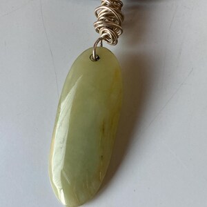 May include: A green jade pendant with a silver setting and a gold wire wrap. The pendant is oval shaped and has a smooth, polished surface.