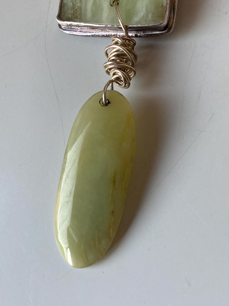 May include: A green jade pendant with a silver setting and a gold wire wrap. The pendant is oval shaped and has a smooth, polished surface.
