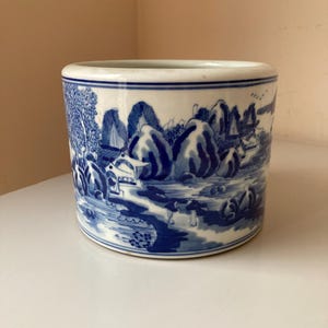 Peut inclure: Un pot de fleurs cylindrique en céramique blanche avec un motif de paysage bleu et blanc. Le pot a une forme ronde et une large ouverture.