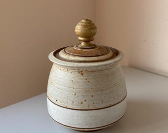 Puede incluir: Un tarro de cerámica blanco con tapa marrón y pomo de madera. El tarro tiene una banda blanca alrededor del medio y motas marrones por todas partes.