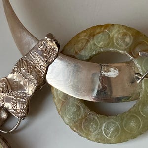 May include: A silver necklace with a green jade pendant. The pendant is a circle with a carved design. The necklace is made of a silver chain with a silver clasp.