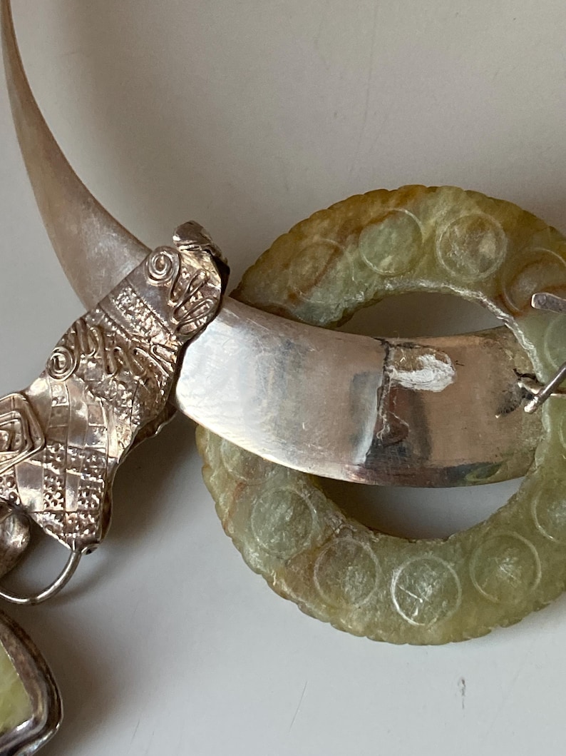 May include: A silver necklace with a green jade pendant. The pendant is a circle with a carved design. The necklace is made of a silver chain with a silver clasp.