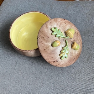 Könnte beinhalten: Eine Keramik-Schale mit Deckel in brauner und grüner Glasur. Der Deckel zeigt ein Design mit drei Eicheln und zwei Eichenblättern.