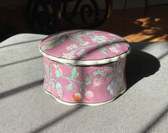May include: A small, round, pink porcelain trinket box with a floral design. The lid has a white rim and the box has a scalloped edge.