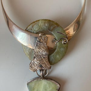May include: A silver necklace with a jade pendant. The pendant is a carved bird with intricate details. The necklace is a wide band with a jade circle attached to it.