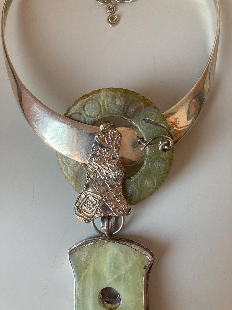 May include: A silver necklace with a jade pendant. The pendant is a carved bird with intricate details. The necklace is a wide band with a jade circle attached to it.