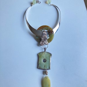 May include: A silver choker necklace with a green stone pendant. The pendant is made up of three pieces: a round green stone with a silver ring around it, a rectangular green stone with a silver frame, and a teardrop-shaped green stone.