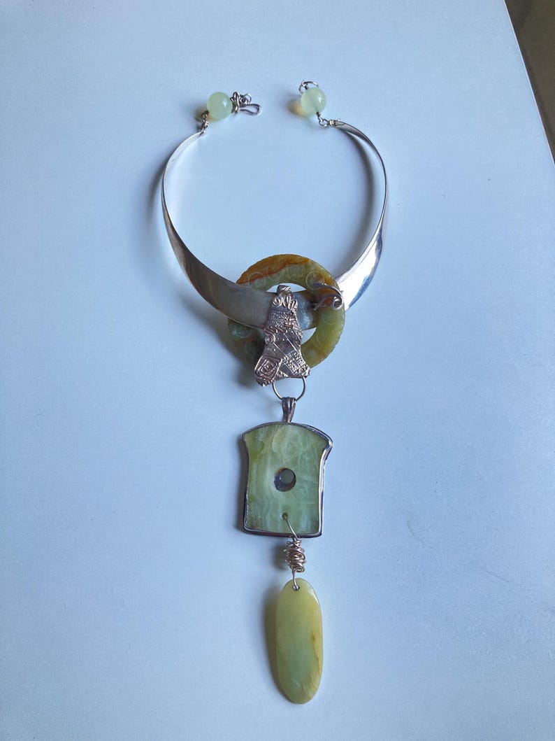 May include: A silver choker necklace with a green stone pendant. The pendant is made up of three pieces: a round green stone with a silver ring around it, a rectangular green stone with a silver frame, and a teardrop-shaped green stone.