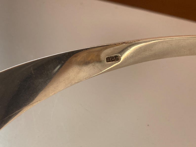 May include: Close-up of a silver bracelet with the number "925" stamped on the inside. The bracelet is curved and has a smooth, polished finish.