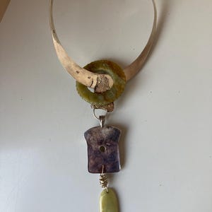 May include: A silver necklace with a green stone pendant. The pendant is a large, round stone with a smaller, round stone in the center. The necklace is attached to a silver chain with a silver clasp. The necklace is on a white surface.