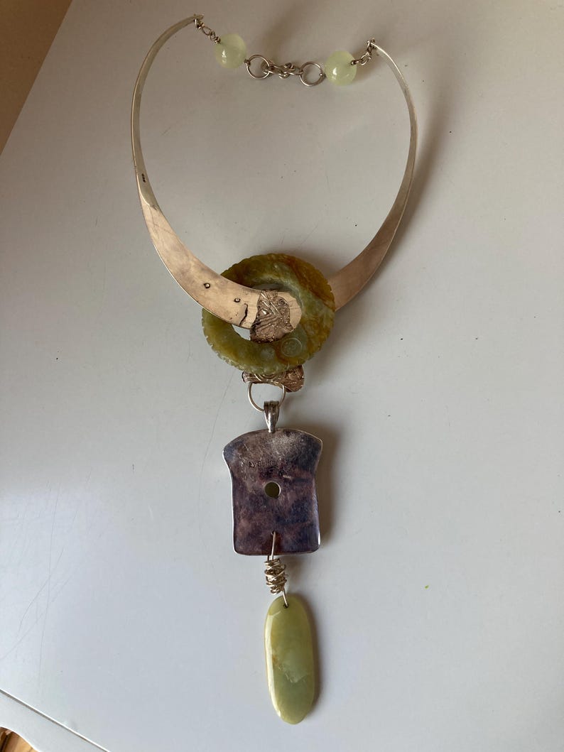 May include: A silver necklace with a green stone pendant. The pendant is a large, round stone with a smaller, round stone in the center. The necklace is attached to a silver chain with a silver clasp. The necklace is on a white surface.
