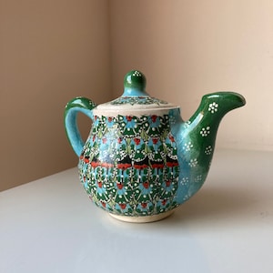 May include: A hand-painted ceramic teapot with a lid, handle, and spout. The teapot features a vibrant pattern of green, blue, red, and white floral motifs. The handle and spout are green, and the lid is white with a green knob.