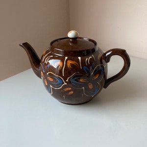 May include: A brown teapot with a white knob on the lid. The teapot is decorated with blue and orange floral designs.