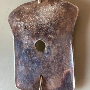 May include: A silver pendant with a textured surface and a hole in the center. The pendant is marked "by Shirley" and "Sterling".