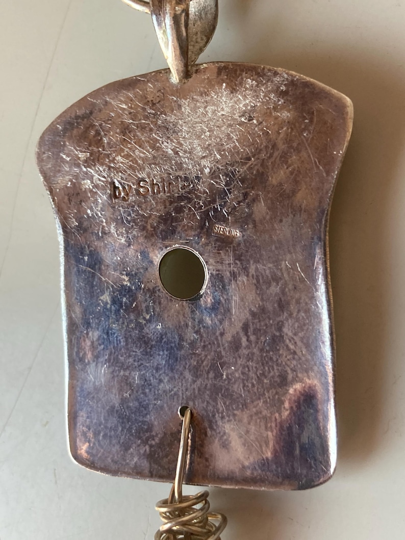 May include: A silver pendant with a textured surface and a hole in the center. The pendant is marked "by Shirley" and "Sterling".