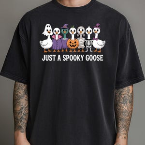 Op de afbeelding: Zwart T-shirt met een Halloween-thema. Het ontwerp toont zeven cartoon ganzen verkleed in enge kostuums, waaronder een spook, vampier en mummie. De tekst "JUST A SPOOKY GOOSE" staat eronder.
