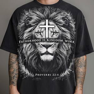 Peut inclure: Un t-shirt noir à manches courtes orné d'un grand graphique détaillé représentant une tête de lion avec une croix blanche entre les yeux. Le texte "FATHERHOOD IS KINGDOM WORK" est imprimé au centre, avec "PROVERBS 22:6" inscrit en bas.