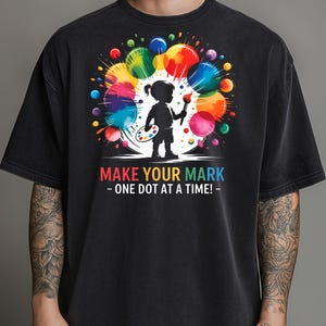 May include: Black t-shirt featuring a colorful graphic of a child artist with a paintbrush and palette, surrounded by vibrant paint splatters. Text reads "MAKE YOUR MARK - ONE DOT AT A TIME!"