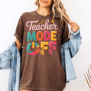 Peut inclure: Un t-shirt marron avec le texte "Teacher Mode Off" en lettres color&eacute;es. Le texte est entour&eacute; de palmiers et d'un soleil.