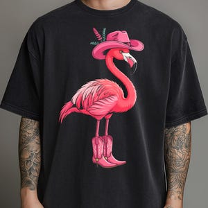 May include: Black t-shirt featuring a pink flamingo wearing a cowboy hat and boots. The flamingo illustration has a whimsical design, with the bird's feathers and accessories in shades of pink.