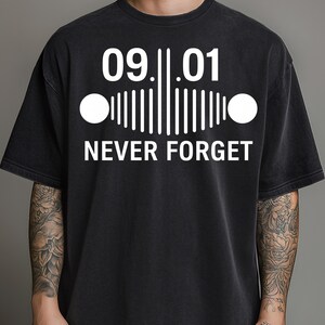 Puede incluir: Camiseta negra con el texto blanco "09.01 NEVER FORGET". El diseño incluye los números "09.01" sobre un gráfico que se asemeja a una parrilla de coche, con dos círculos a cada lado. La camiseta tiene mangas cortas.