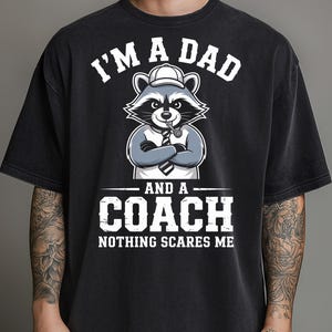 May include: Black t-shirt featuring a cartoon raccoon wearing a hat and tie, with the text "I'M A DAD AND A COACH NOTHING SCARES ME" in white. The raccoon has crossed arms and a whistle in its mouth.