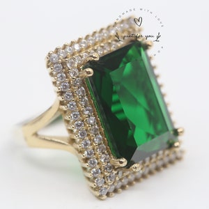May include: A gold ring with a large emerald green gemstone surrounded by small clear stones. The ring is set in a square shape with a double row of stones around the gemstone.