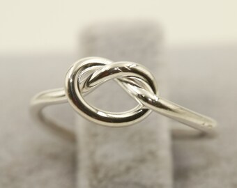 May include: A silver-colored ring featuring a knot design. The ring is made of a shiny, reflective metal and sits on a gray surface. The knot is the focal point, with the band extending to the sides. A simple, elegant piece of jewelry.