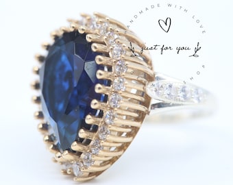 May include: A gold ring with a large, dark blue gemstone surrounded by smaller, clear gemstones. The ring is set in a gold band with a silver band on the side. The ring is inscribed with the words "Handmade with Love, just for you".