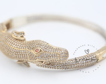 May include: A gold alligator bracelet with a red stone eye and a silver band. The bracelet is covered in small, clear stones. The bracelet is handmade with love, just for you.