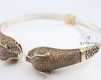 May include: A silver and gold bangle bracelet with two panther heads on either end. The panther heads are covered in small brown stones and have green eyes. The bracelet is engraved with the words "Handmade with Love, just for you".