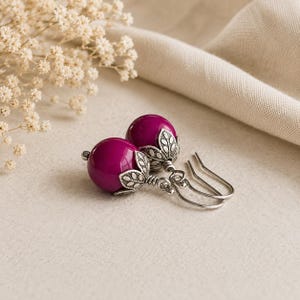 May include: A pair of drop earrings featuring round, deep magenta beads. Each bead is capped with an antique silver-toned metal finding shaped like a small, stylised leaf-patterned calyx. The earrings are attached to simple silver-toned metal hooks.