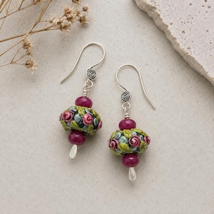 Puede incluir: Un par de pendientes colgantes con ganchos de plata y un detalle en espiral. Cada pendiente tiene una cuenta central redonda decorada con un patrón floral de rosas rosadas y hojas verdes, flanqueada por dos cuentas magenta facetadas.