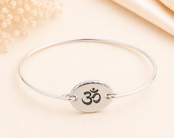 May include: A silver bangle bracelet featuring a central oval charm with a hammered edge and a black engraved Om symbol. The thin, polished metal band connects to the charm on both sides, creating a continuous circular design for the wrist.