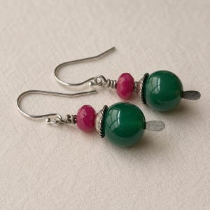May include: A pair of drop earrings featuring a large, smooth, dark green spherical bead paired with a smaller, faceted, bright pink bead. The beads are connected by silver-toned wire components and finished with simple silver-toned ear hooks.
