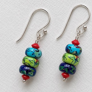 Può includere: Un paio di orecchini pendenti con ganci color argento e una disposizione verticale di perline. Ogni orecchino include una piccola perlina rossa in alto e in basso, con tre perline più grandi a motivi turchese, verde lime e blu scuro.