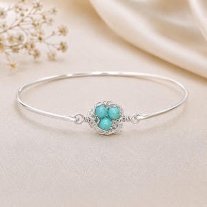 Puede incluir: Una pulsera rígida de plata con un diseño central de nido hecho con alambre. El nido contiene tres pequeñas cuentas redondas de color turquesa brillante. La banda metálica es fina, lisa y circular, conectada al elemento central por ambos lados.