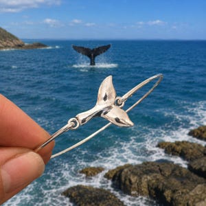 May include: A silver-toned metal bangle bracelet featuring a central whale tail charm. The bracelet has a thin, circular band that connects to the sides of the smooth, polished whale tail ornament, creating a continuous, lightweight jewellery piece.