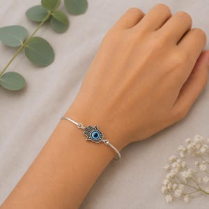 May include: A silver-toned metal bangle bracelet featuring a central Hamsa hand charm. The charm is detailed with a small, circular blue and white evil eye bead at its centre, set against a dark, oxidised silver background with intricate line work.