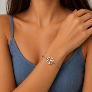 May include: A silver-toned metal bracelet featuring a circular charm with a cut-out world map design. The bracelet consists of a thin, smooth band that connects to the central globe pendant, resting on the wrist of a person wearing a blue tank top.